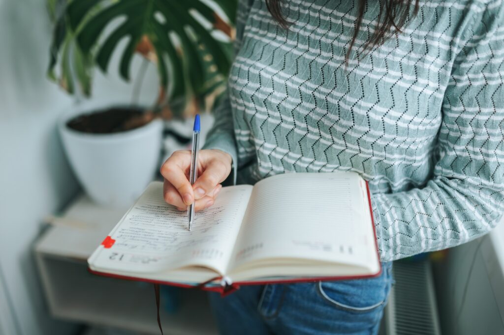 Unrecognizable girl woman standing makes important notes in workbook, pen on paper.